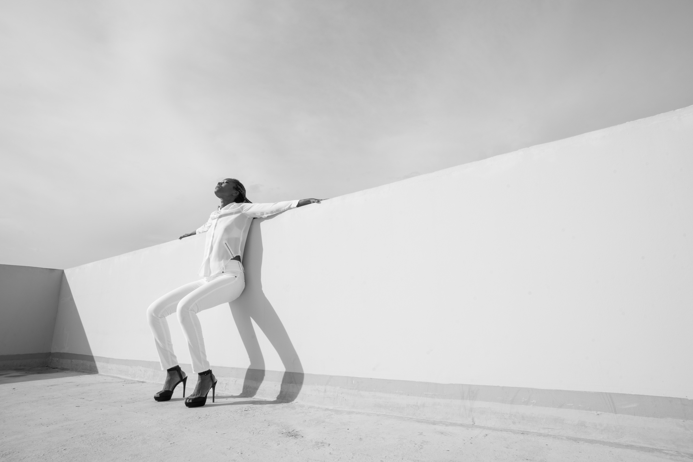 Fotografía en blanco y negro de una mujer vestida con camisa y pantalones blancos y zapatos negros de tacón alto, apoyada contra una pared blanca en una azotea. Mantiene los brazos extendidos a ambos lados y el rostro inclinado hacia el cielo. La luz del sol proyecta una sombra alargada de su figura sobre la pared, mientras las líneas diagonales de la arquitectura dominan la composición.