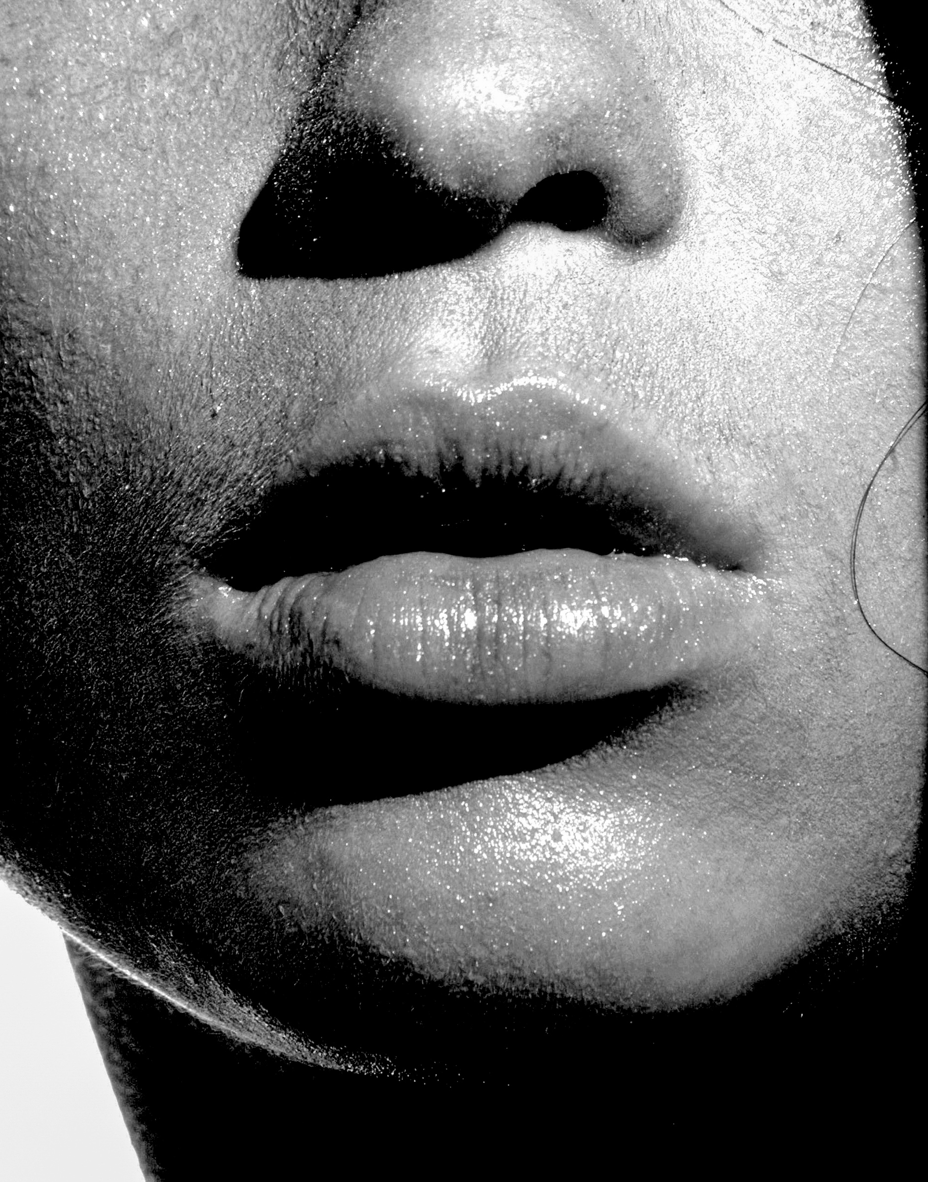 Fotografía en blanco y negro de un primerísimo plano de la parte inferior de un rostro. El encuadre muestra la nariz, los labios ligeramente entreabiertos y el mentón, dejando fuera los ojos. La iluminación intensa resalta el brillo de los labios y la textura de la piel, mientras profundas sombras negras acentúan los contornos del rostro.