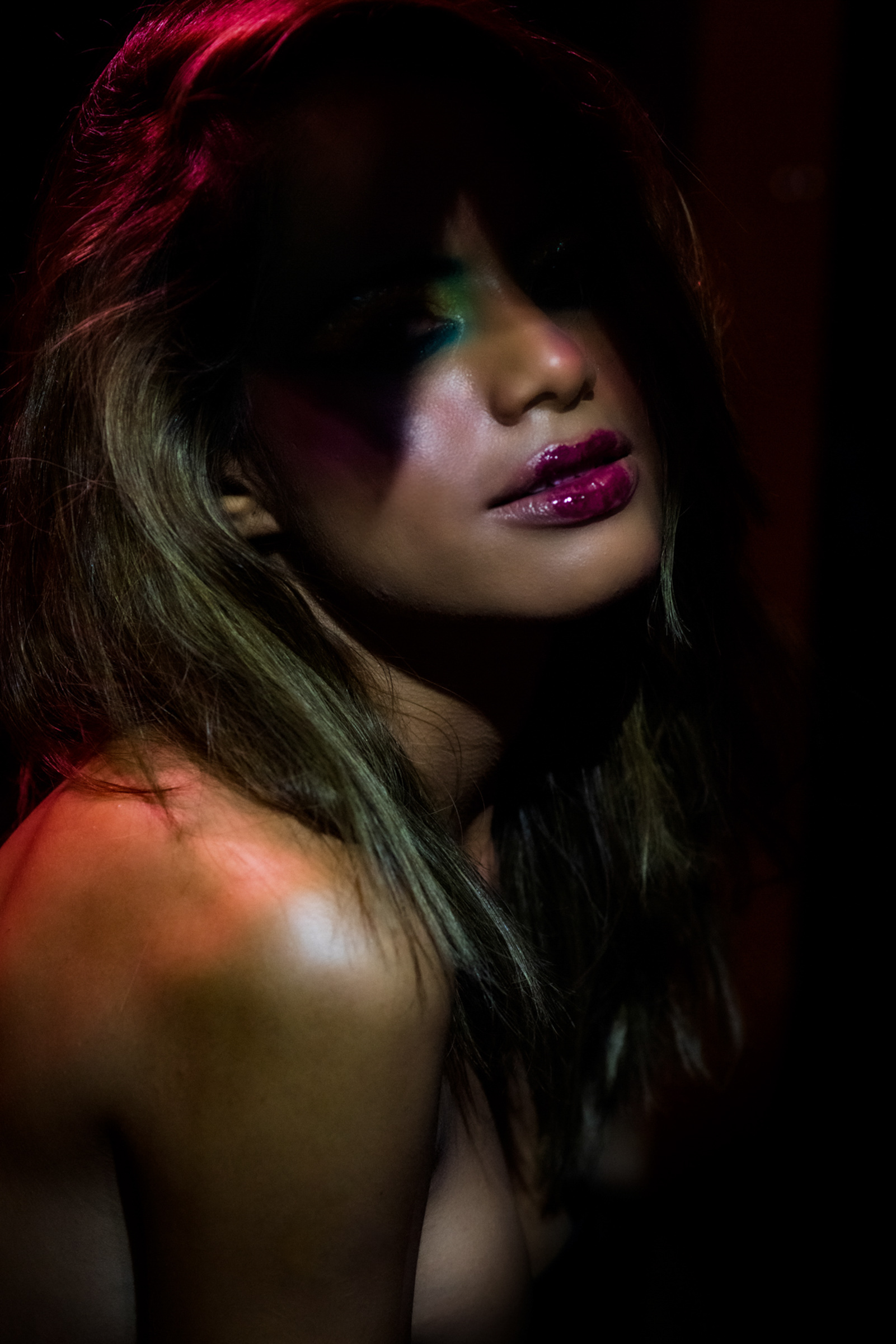 Retrato de una mujer de cabello largo y oscuro, con el rostro ligeramente inclinado y parcialmente oculto por su cabello. Sus labios llevan un intenso brillo fucsia y sus ojos permanecen entre sombras. Luces rojas, verdes y magentas iluminan distintas partes de su rostro, cabello y hombro descubierto, sobre un fondo casi negro.